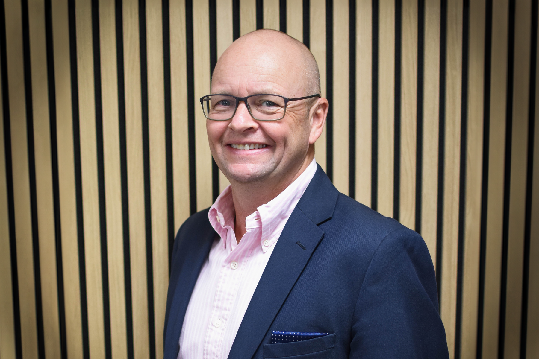 Man with glasses and smart suit stood in front of wood panelled background.